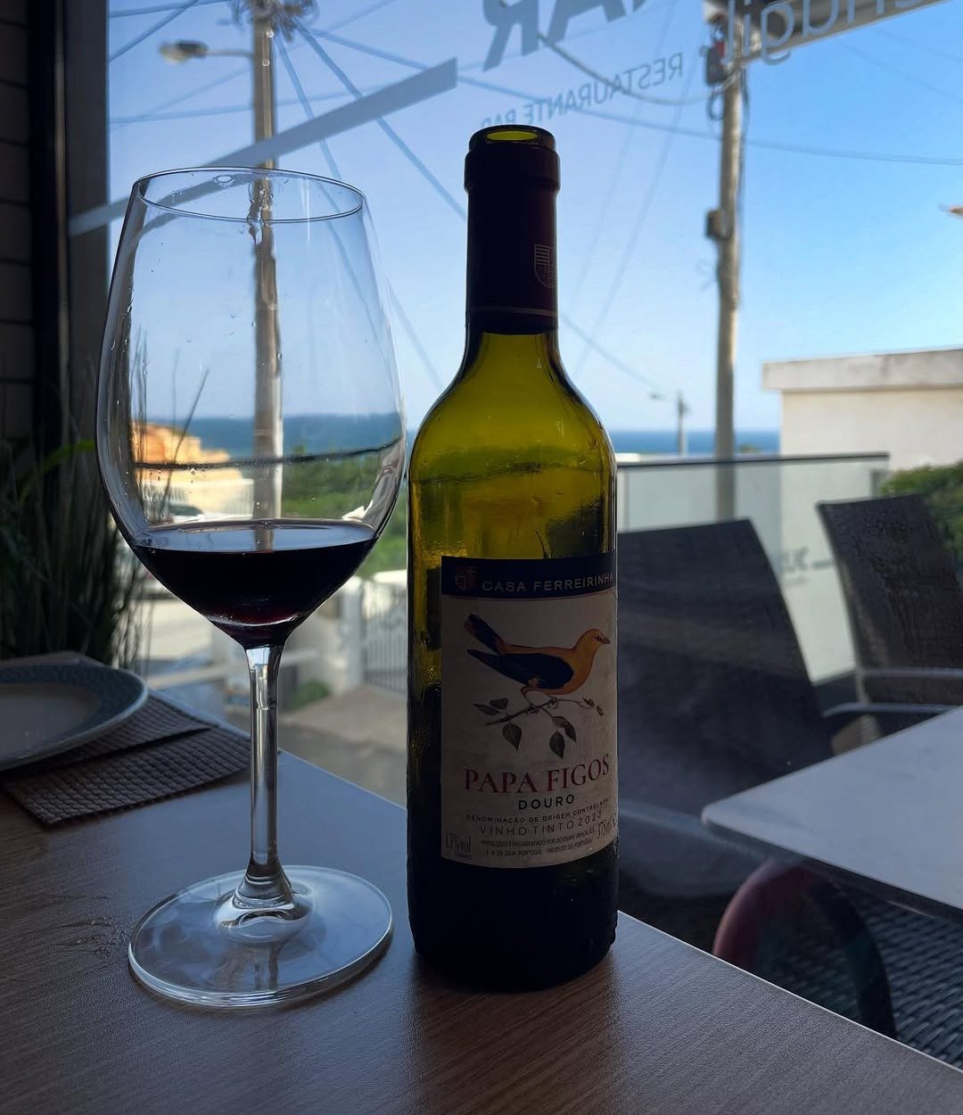 Copo e garrafa de vinho tinto numa mesa junto ao vidro, com a esplanada e o mar ao fundo