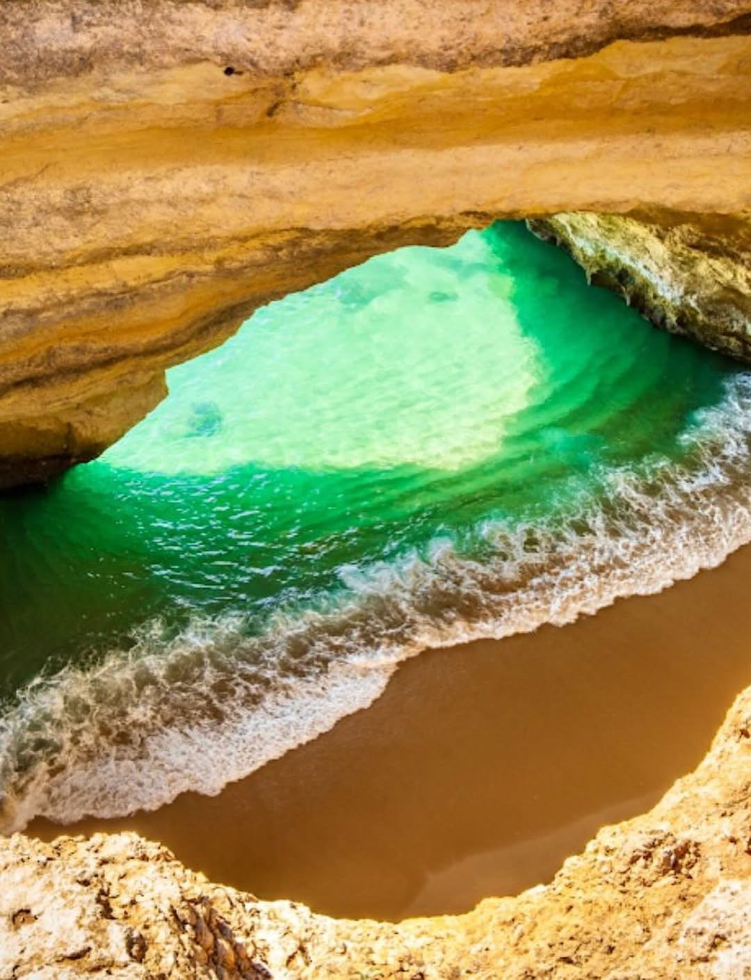 Vista de cima de uma abertura na falésia, com mar verde e areia dourada